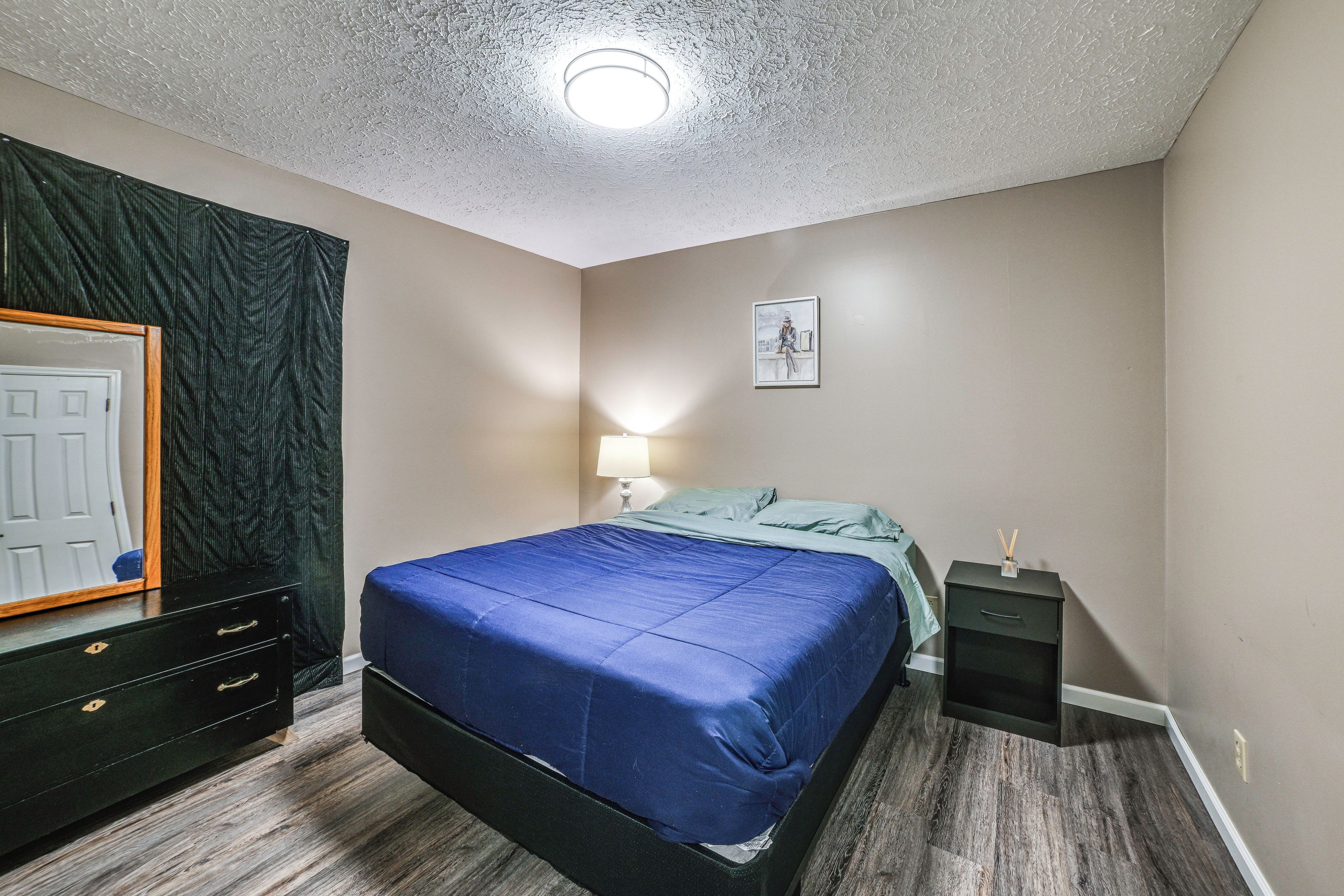 A bedroom with a bed, a nightstand, a lamp, a framed picture on the wall, and a mirror with a dark curtain.
