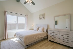 Um quarto com uma cama, cômoda, ventilador de teto, janela com cortinas e um quadro na parede.