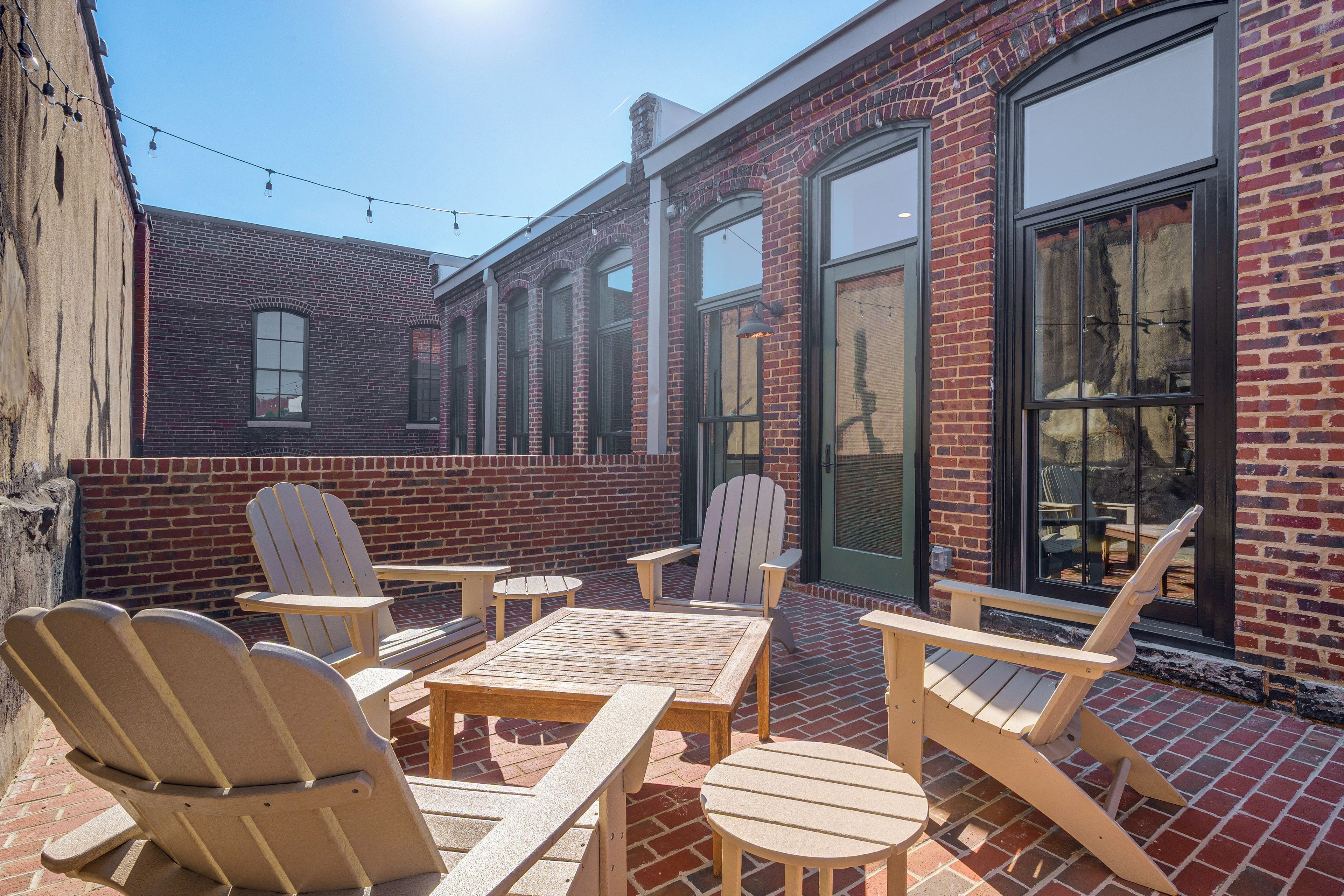 A patio area with Adirondack chairs and tables, a brick building with large windows, and string lights.