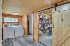 A laundry area with two washing machines and a drying rack, a wooden door leading to a storage room with shelves, and a window with a view of trees.