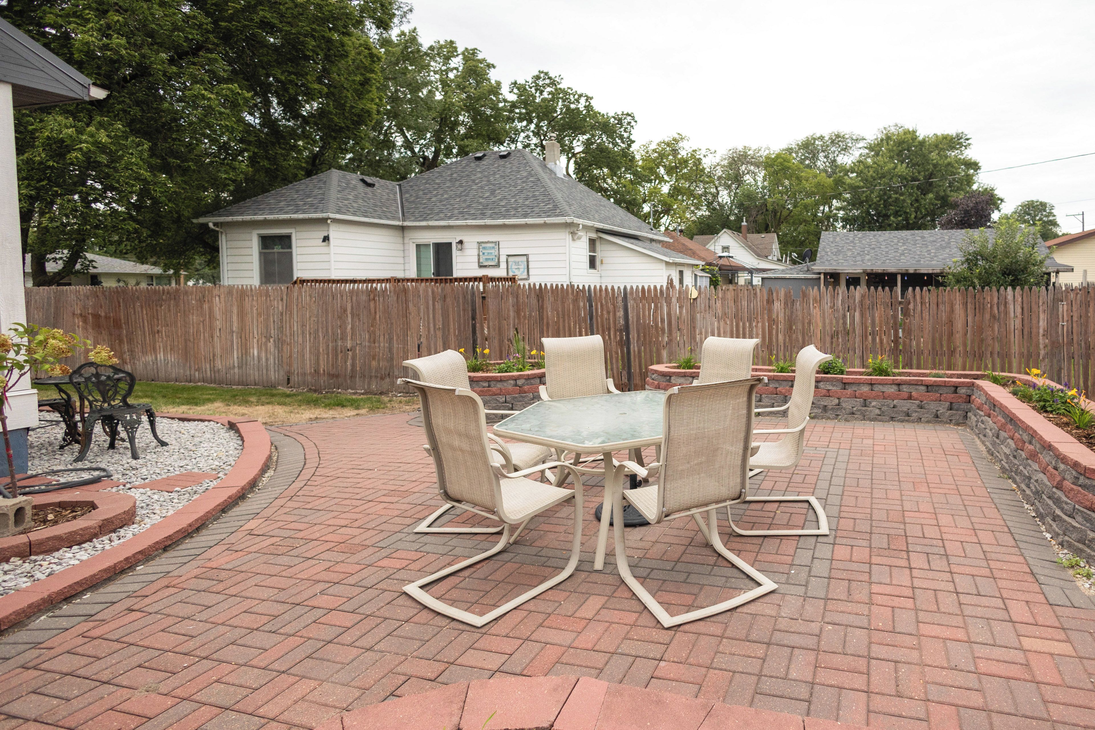 An outdoor patio with a glass-top table and six chairs, surrounded by a brick patio and a wooden fence.