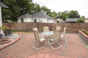 An outdoor patio with a glass-top table and six chairs, surrounded by a brick patio and a wooden fence.