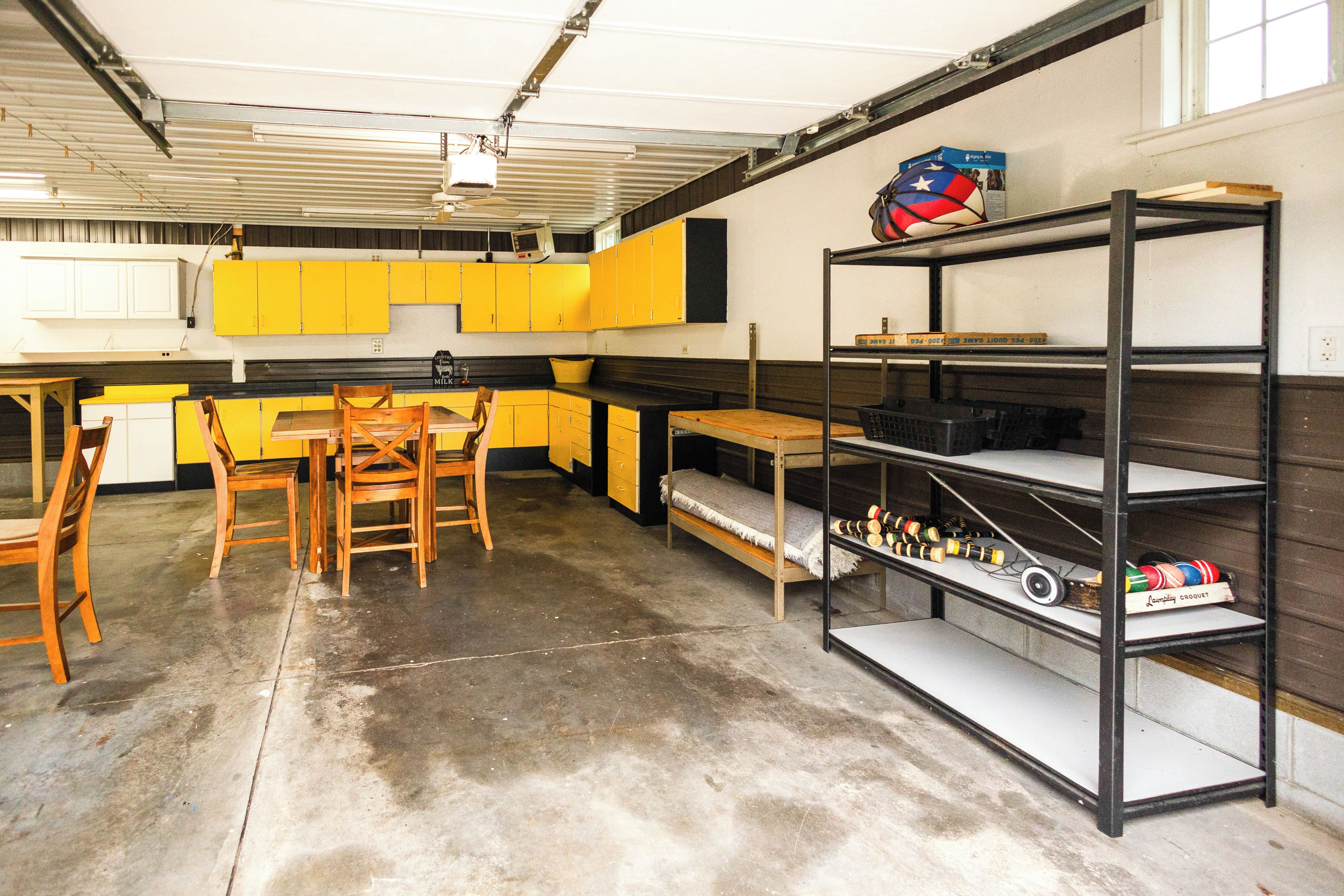 A garage with a kitchen area, wooden chairs, a table, and shelves with various items.