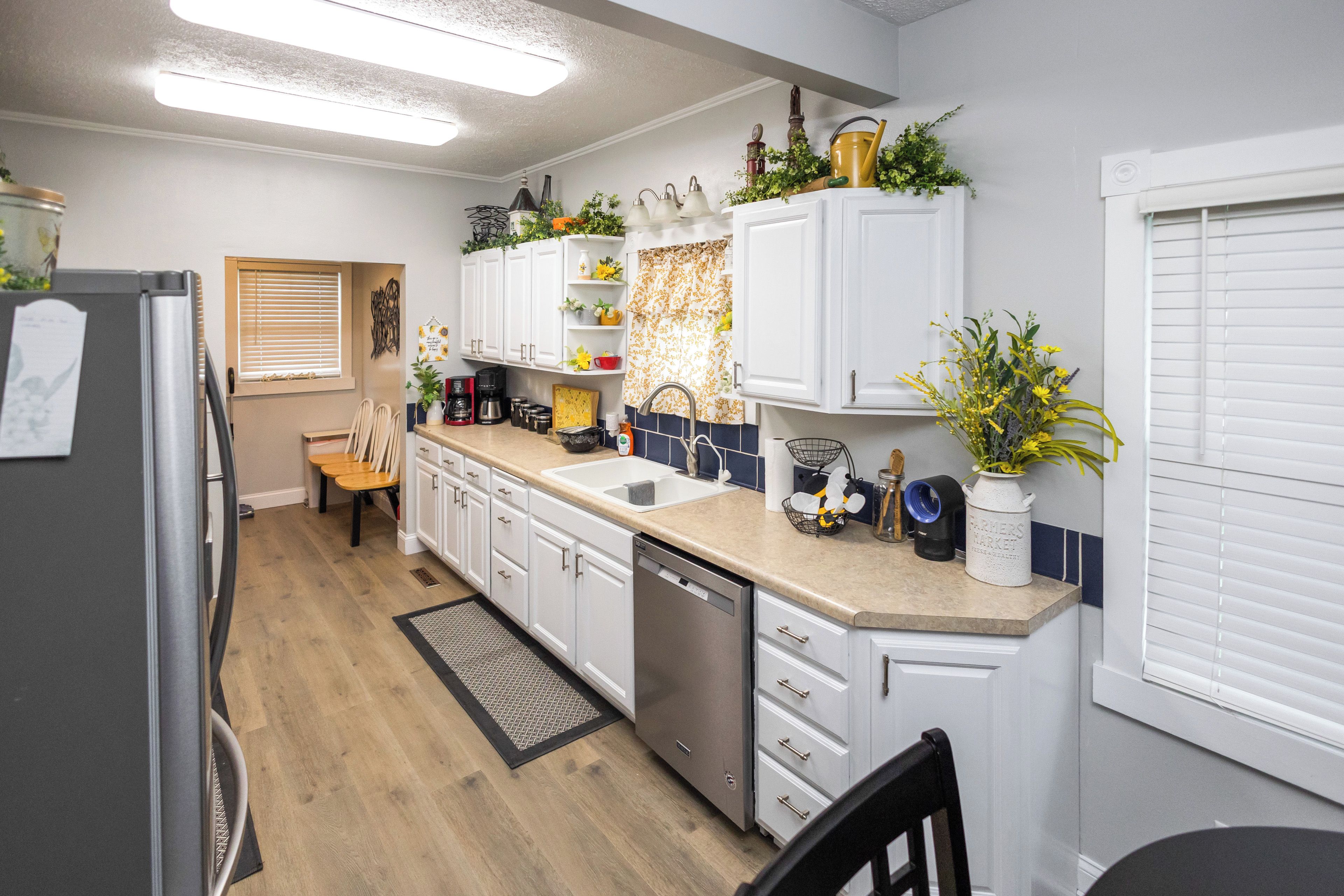 A modern kitchen with white cabinets, a dishwasher, and a sink. There is a window with blinds, a wooden chair, and a small table.