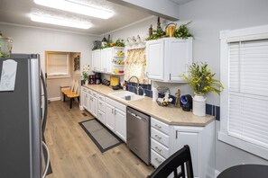 A modern kitchen with white cabinets, a dishwasher, and a sink. There is a window with blinds, a wooden chair, and a small table.