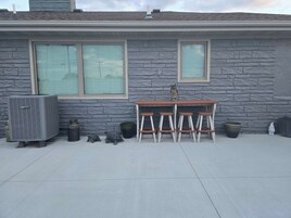 Un patio con pared de piedra, dos ventanas, una mesa y cuatro taburetes, además de unidades de aire acondicionado exteriores.