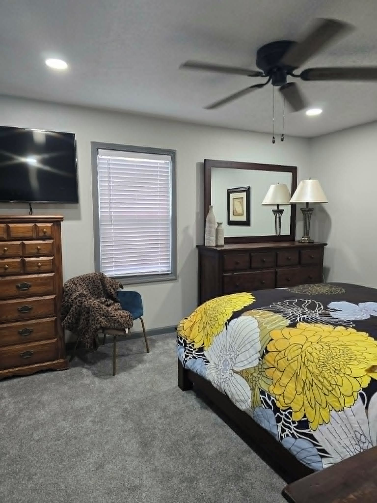 A bedroom with a bed, dresser, television, ceiling fan, window with blinds, and a lamp.