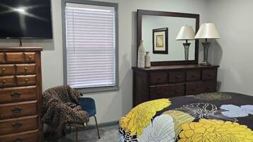A bedroom with a bed, dresser, television, ceiling fan, window with blinds, and a lamp.