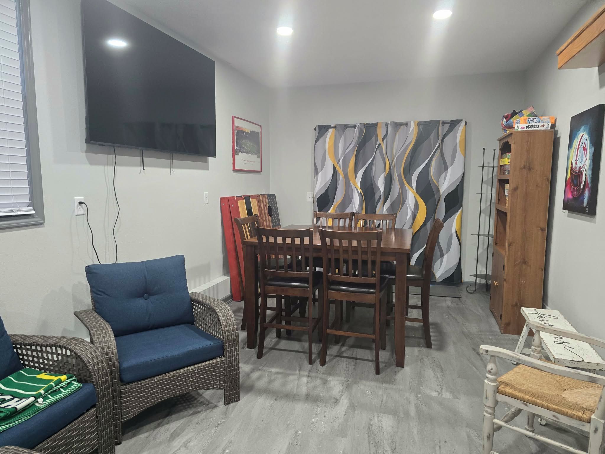 Uma sala de jantar com mesa e cadeiras, uma televisão fixada na parede e um quadro na parede.