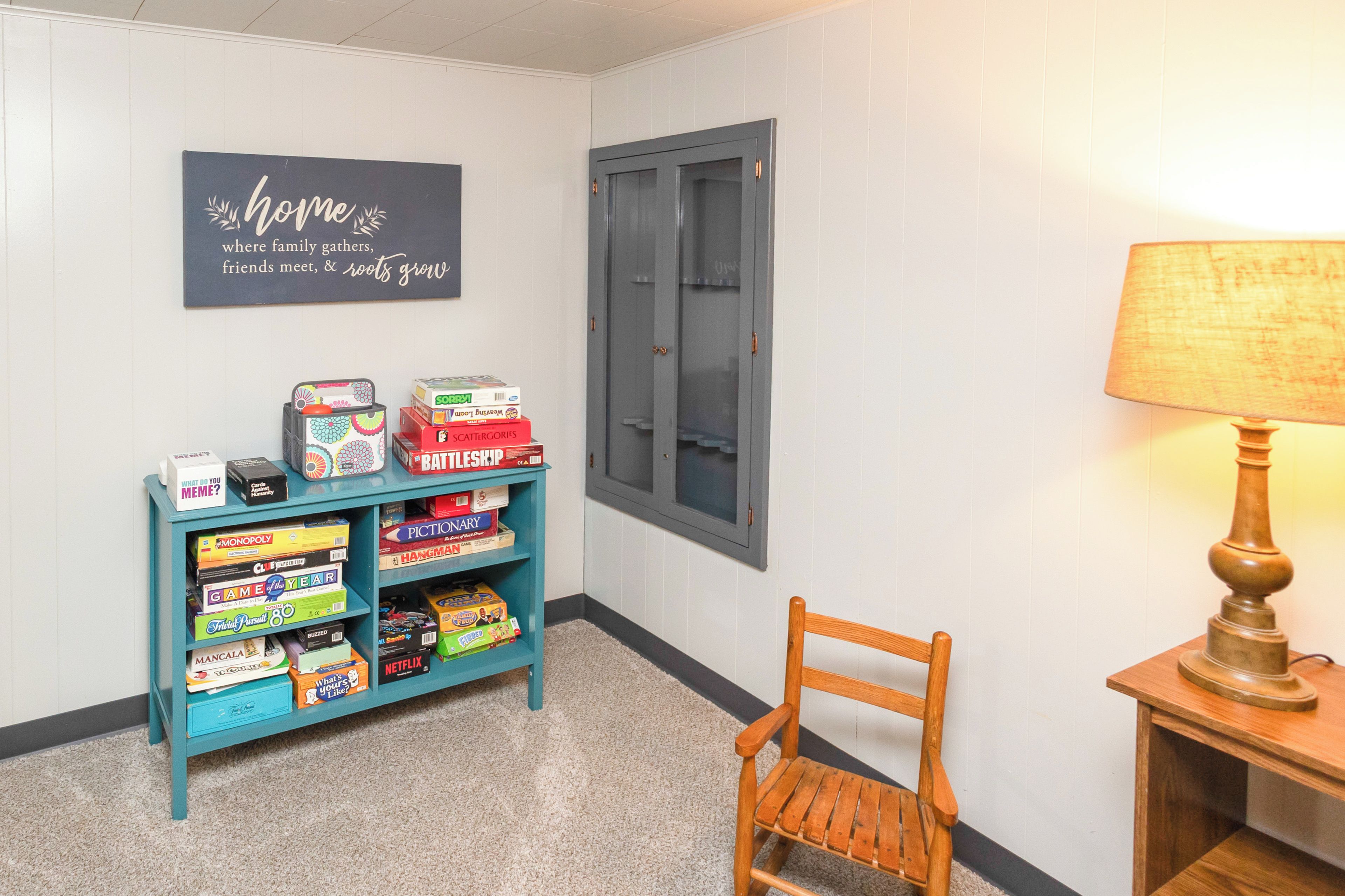 A hallway with a teal shelf displaying board games, a wooden chair, a lamp, and a door with a window.