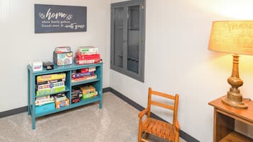 A hallway with a teal shelf displaying board games, a wooden chair, a lamp, and a door with a window.