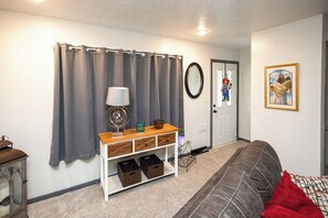 A living room with a grey sofa, a wooden side table, a lamp, a mirror, and a door with a window.