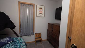 A bedroom with a bed, a dresser, a small bench, a window with curtains, and a wall-mounted TV.