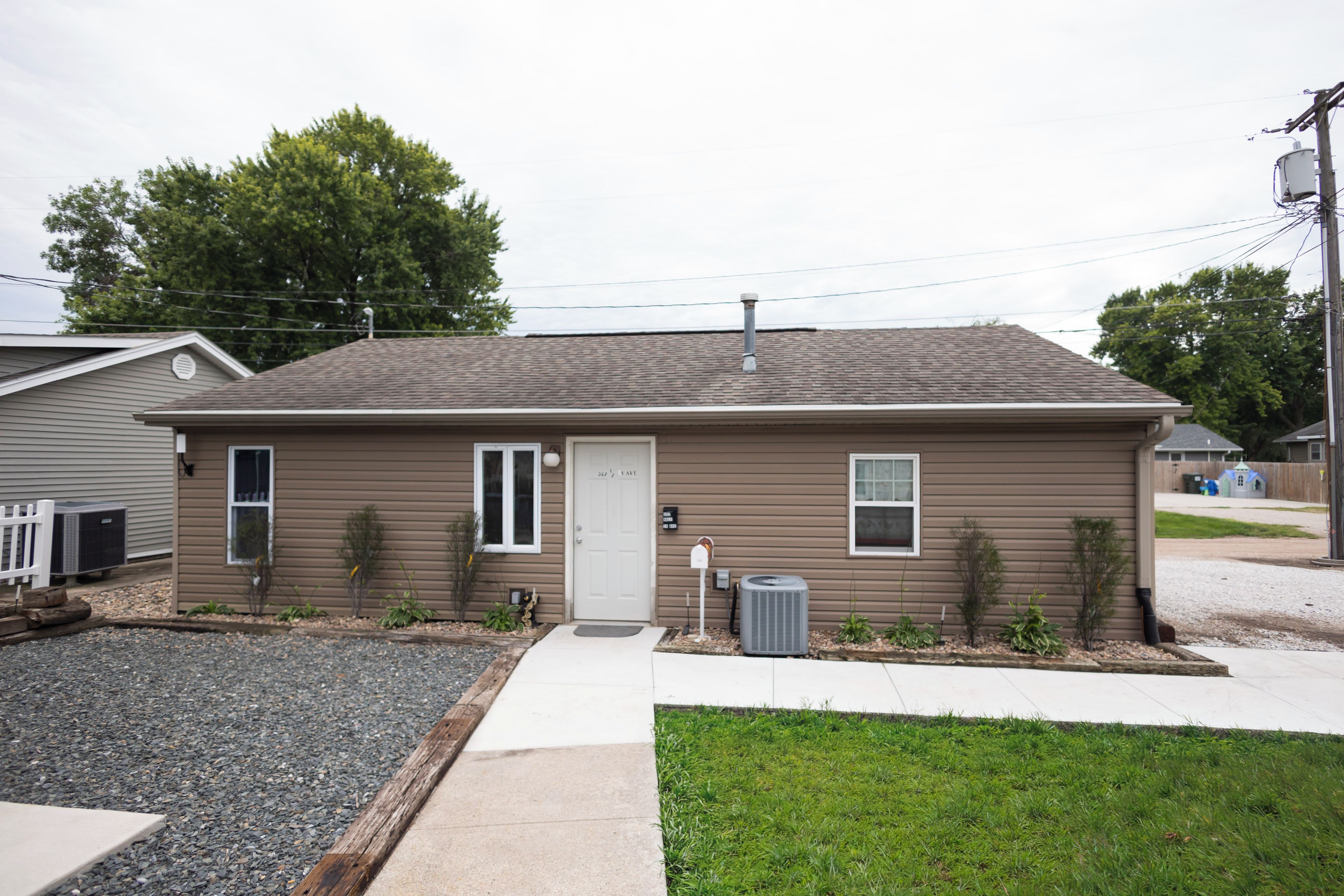 A single-story residential building with a front door, two windows, a small porch area, and a front yard with gravel and grass.