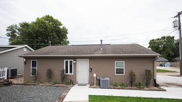 A single-story residential building with a front door, two windows, a small porch area, and a front yard with gravel and grass.