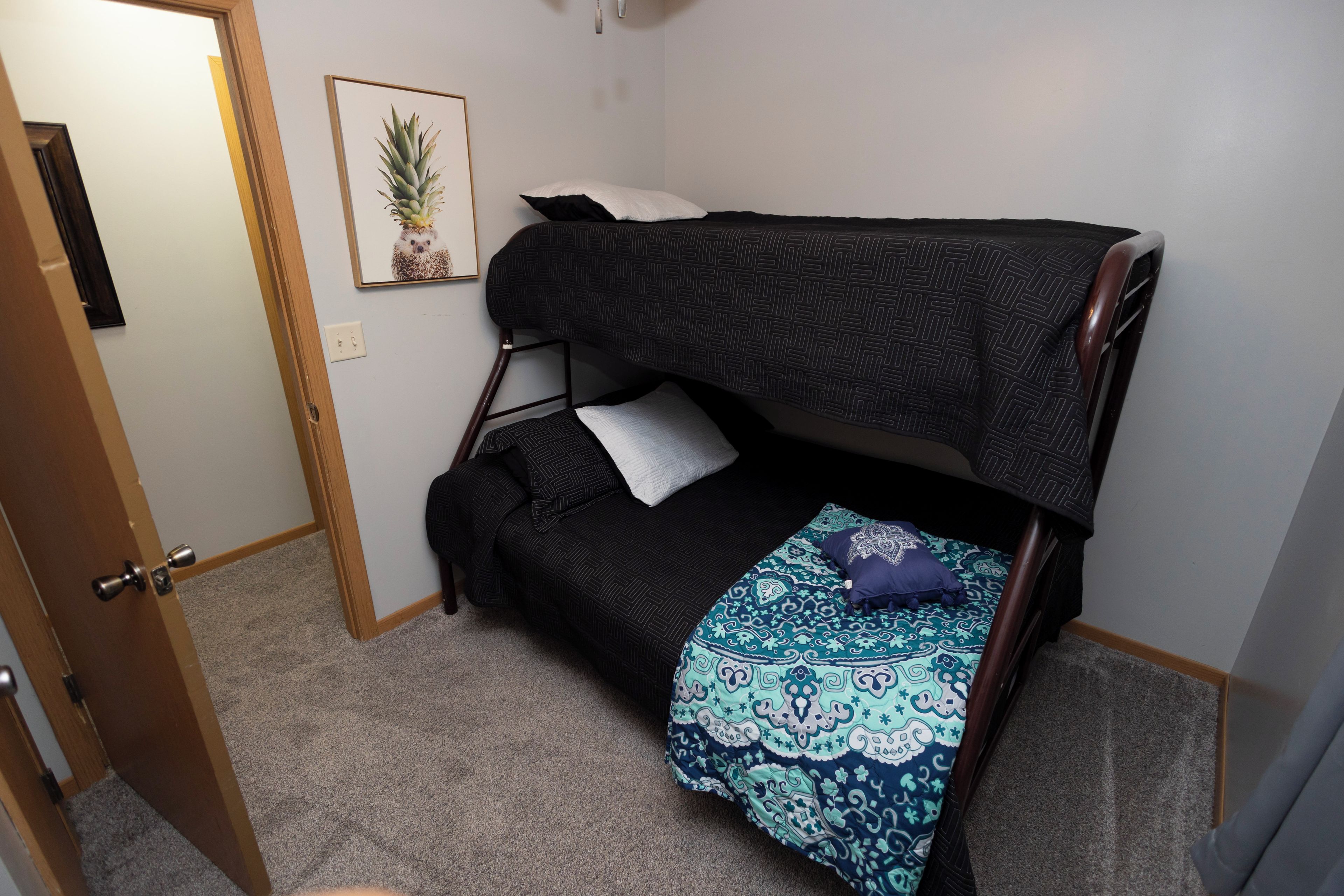 Une petite pièce avec un lit superposé, une porte, un tableau encadré au mur et un sol recouvert de moquette.