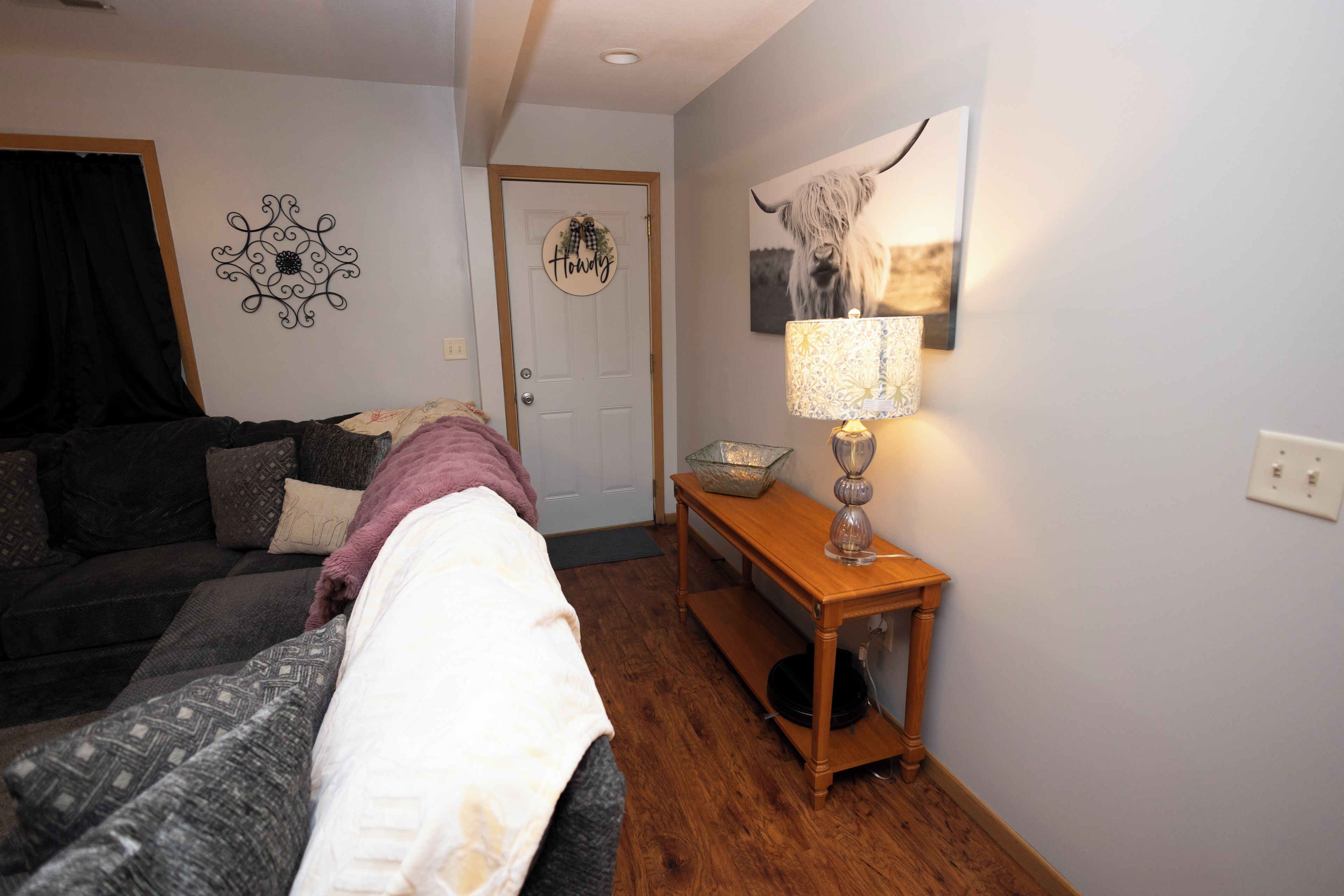 A room with a bed, a wooden table with a lamp, a door with a sign, and a wall decoration.