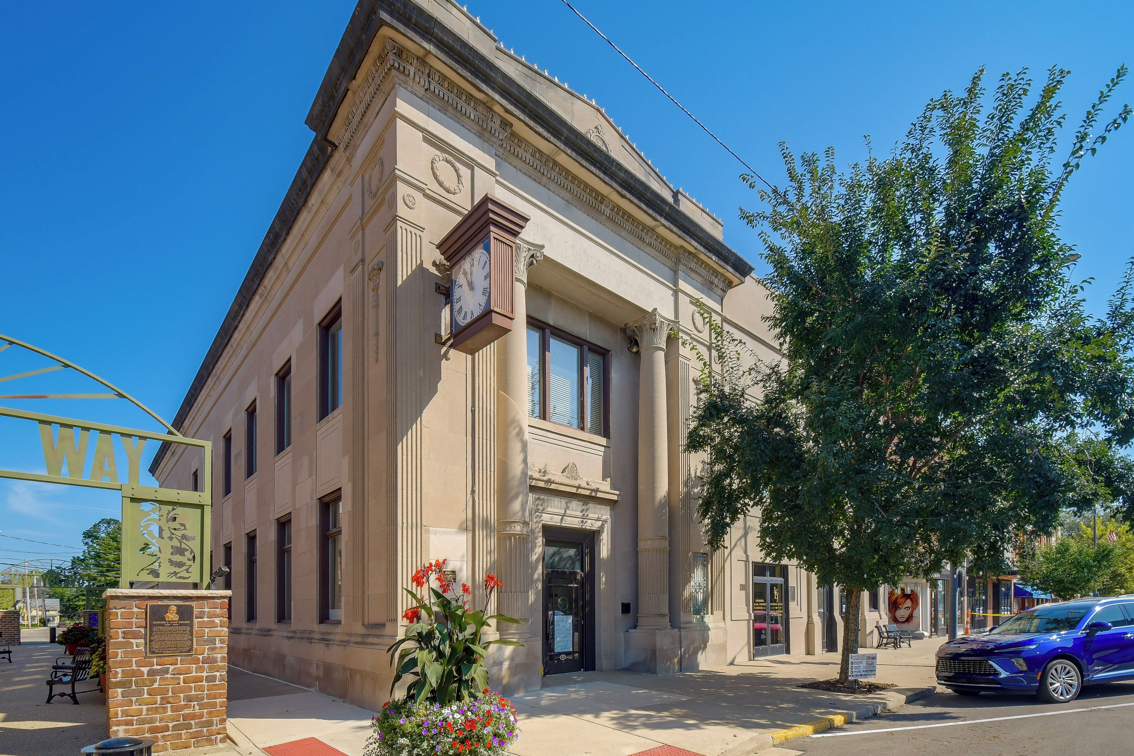 A classical building with a clock, a bench, and a tree in front.