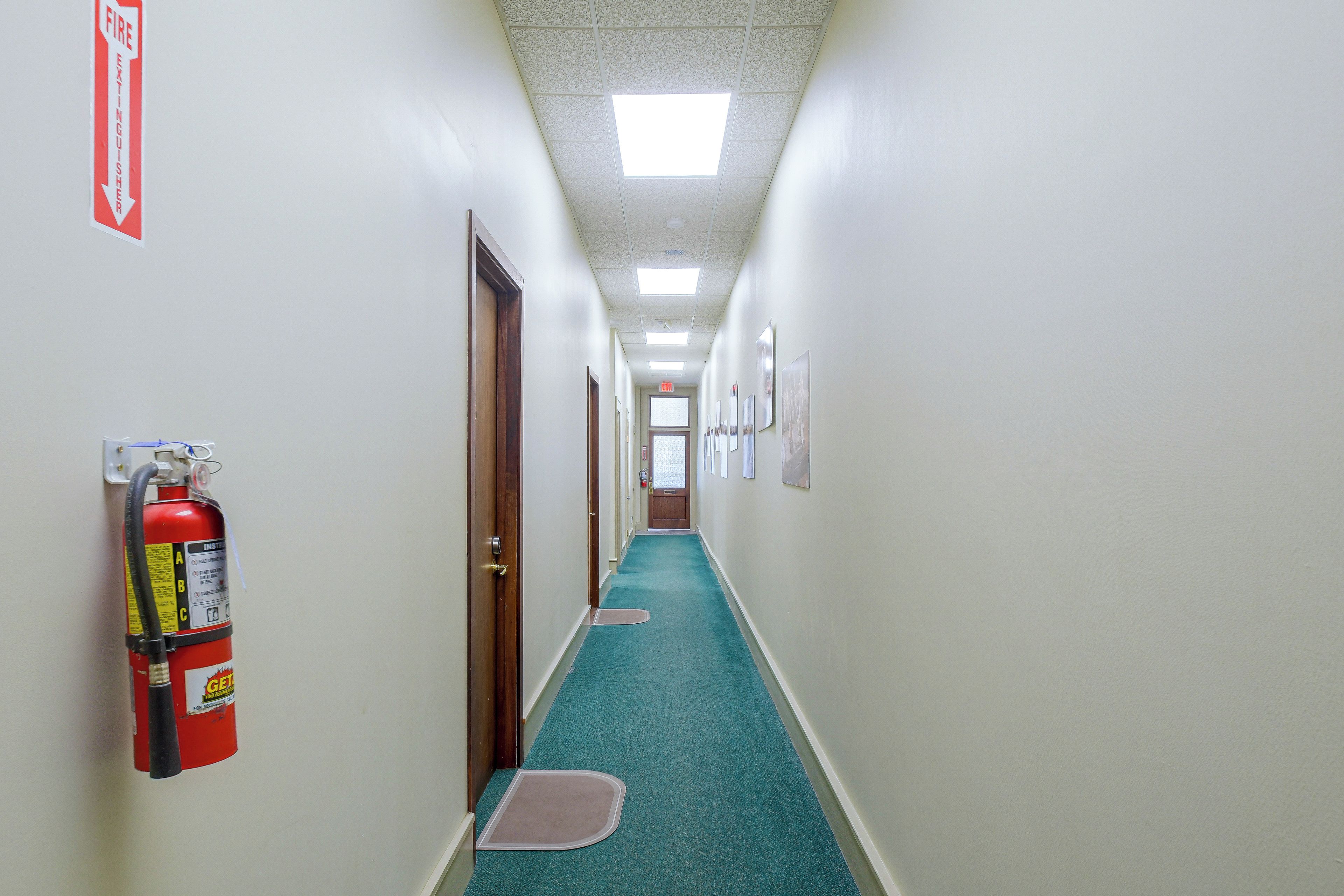 A hallway with a fire extinguisher mounted on the wall, a fire exit sign, and a door at the end.