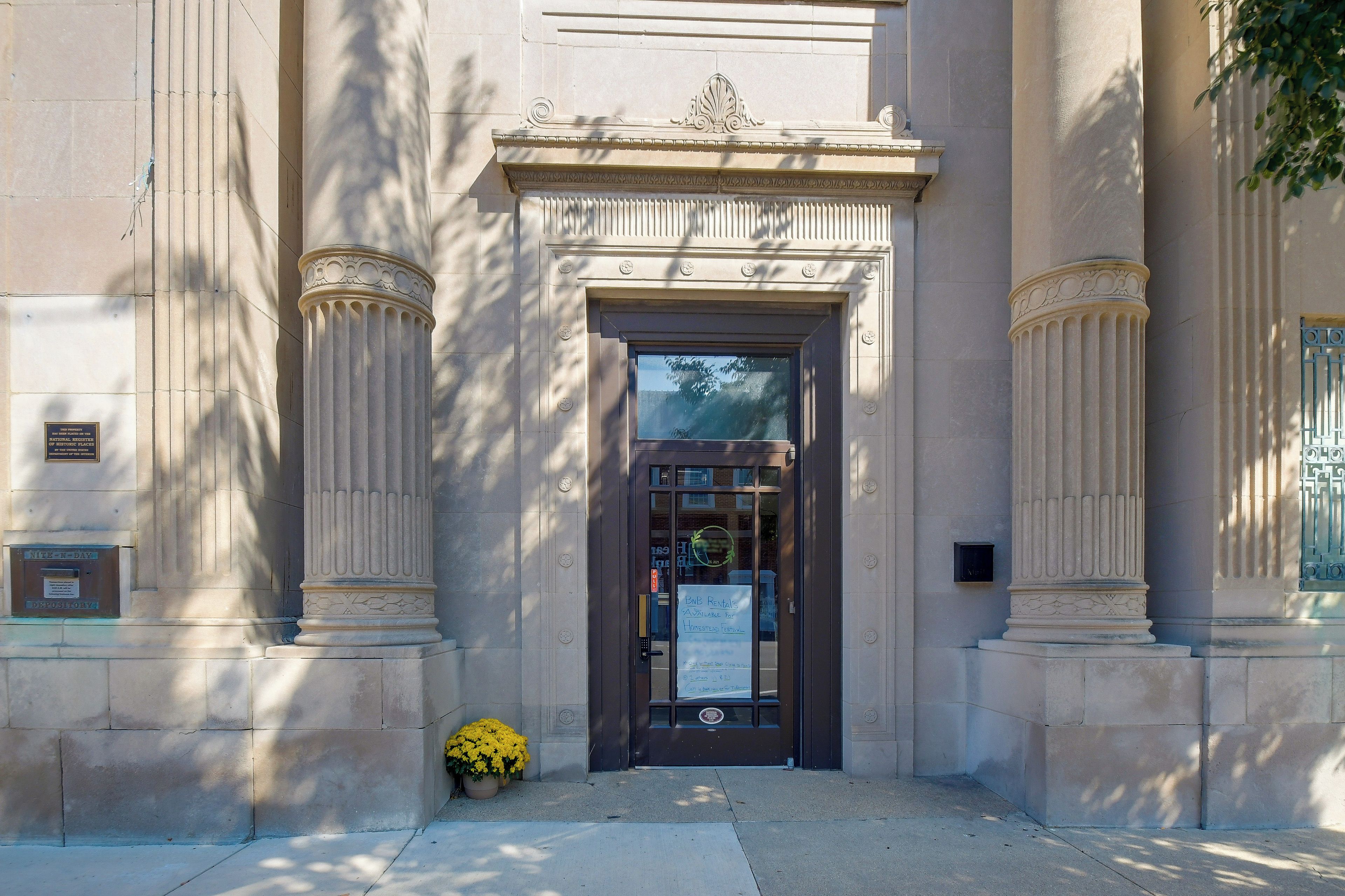 A building entrance with a dark door, flanked by two columns with decorative capitals, and a small potted plant on the ground.