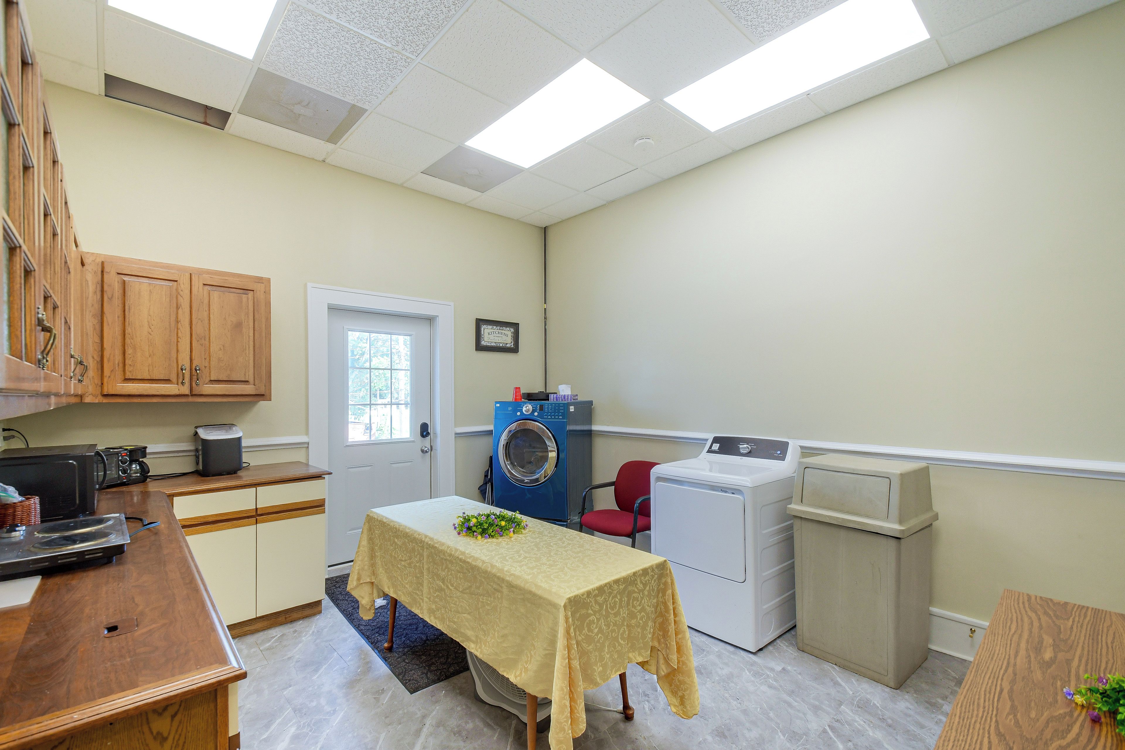 A compact kitchen with a microwave, toaster, and sink. There is a table with a floral arrangement and a red chair.