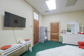 A hotel room with a flat-screen TV mounted on the wall, a wooden door, a white dresser with a mirror, and a green carpet.