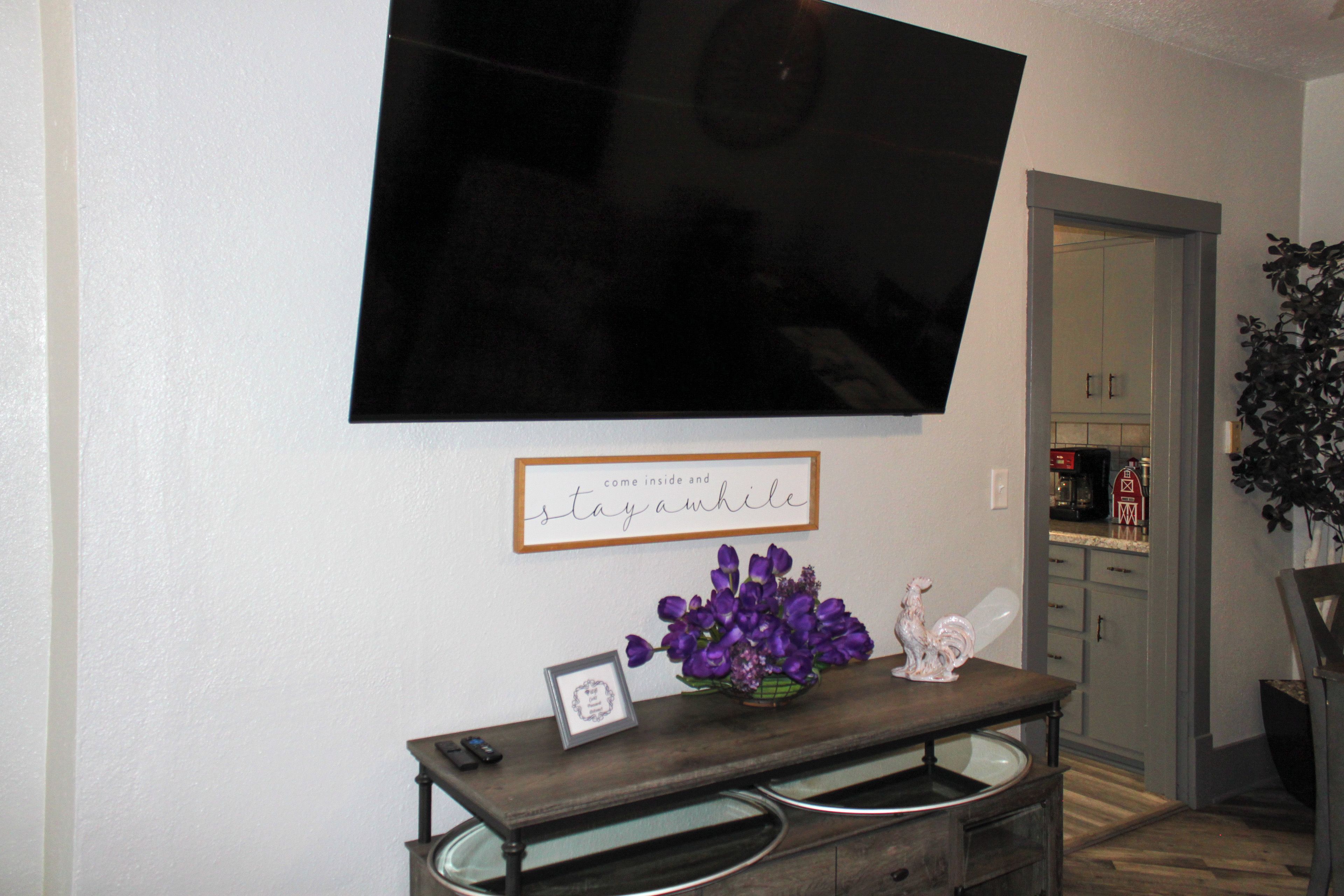 Un salon avec un téléviseur à écran plat fixé au mur, un panneau encadré portant l’inscription « entrez et restez un moment », une petite table sur laquelle est posé un vase rempli de fleurs violettes, et une statue de coq décorative.