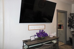 Un salón con un televisor de pantalla plana montado en la pared, un letrero enmarcado con el texto 'come inside and stay awhile', una mesita con un jarrón de flores moradas y una estatua decorativa de gallo.
