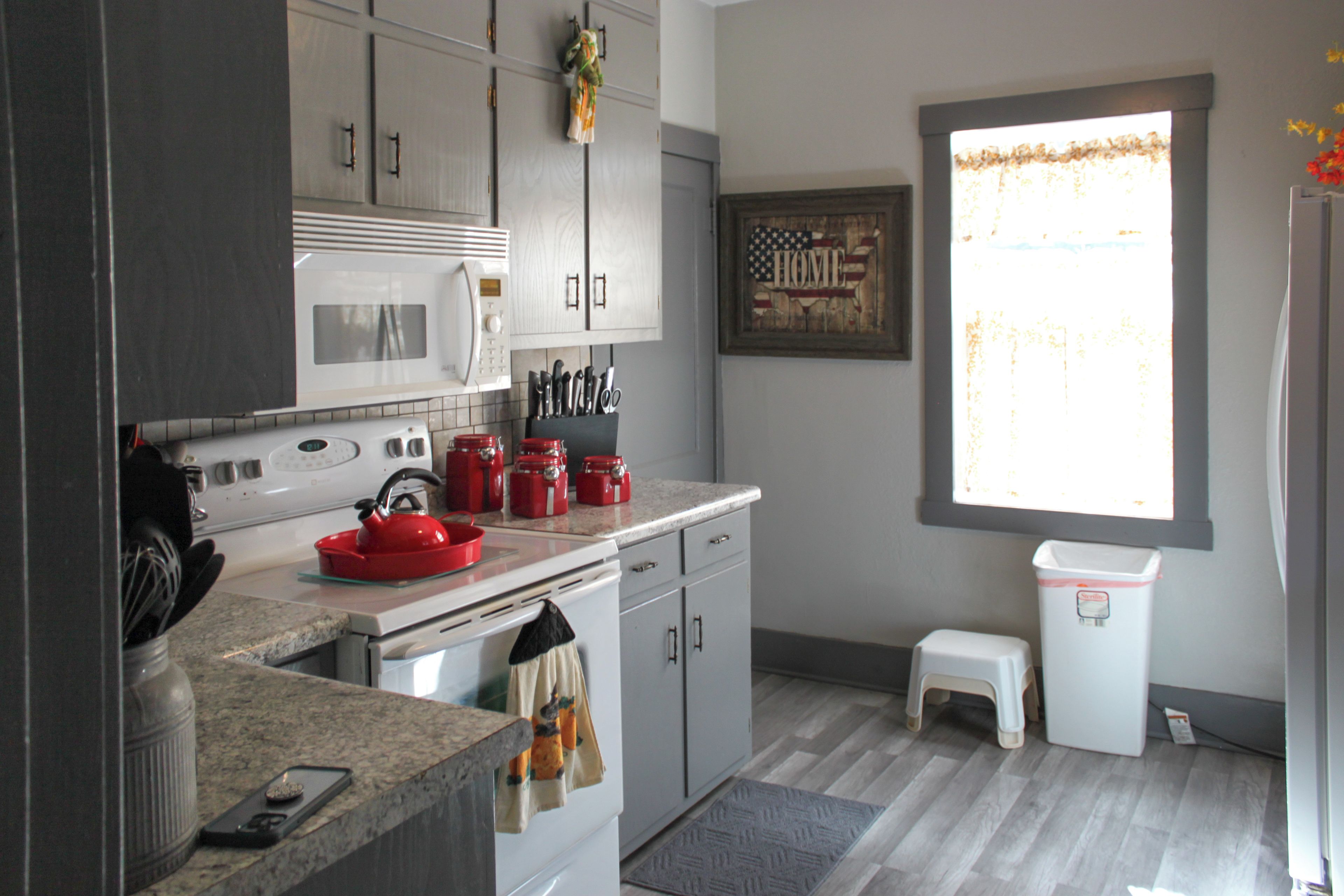A kitchen with a microwave, stove, and red kettle. There is a window with a view of the outside.