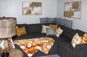 Una sala de estar con un sofá oscuro, varios cojines, un plaid floral y dos cuadros colgados en la pared.