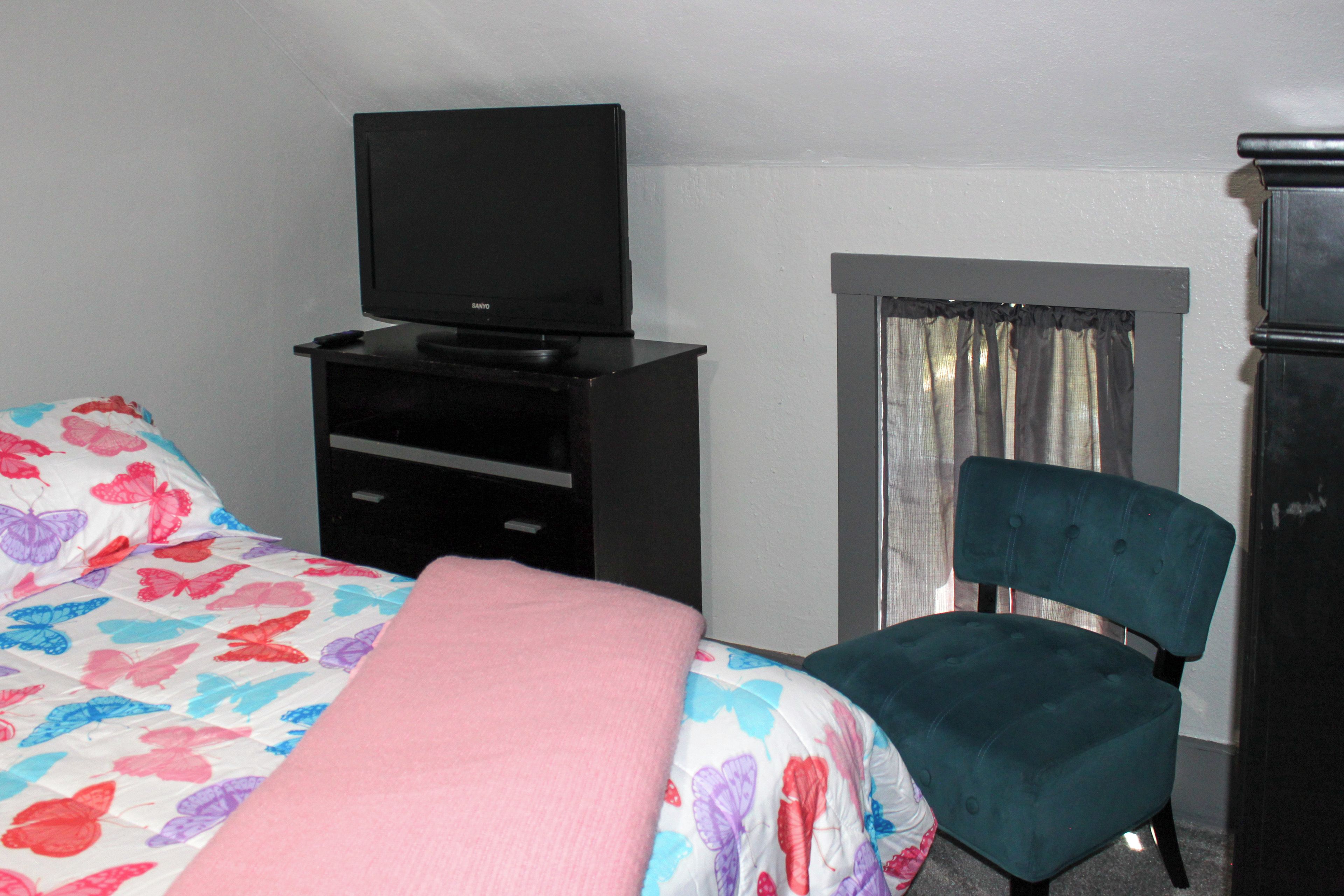 A hotel room with a bed, a chair, a TV on a stand, and a window with curtains.
