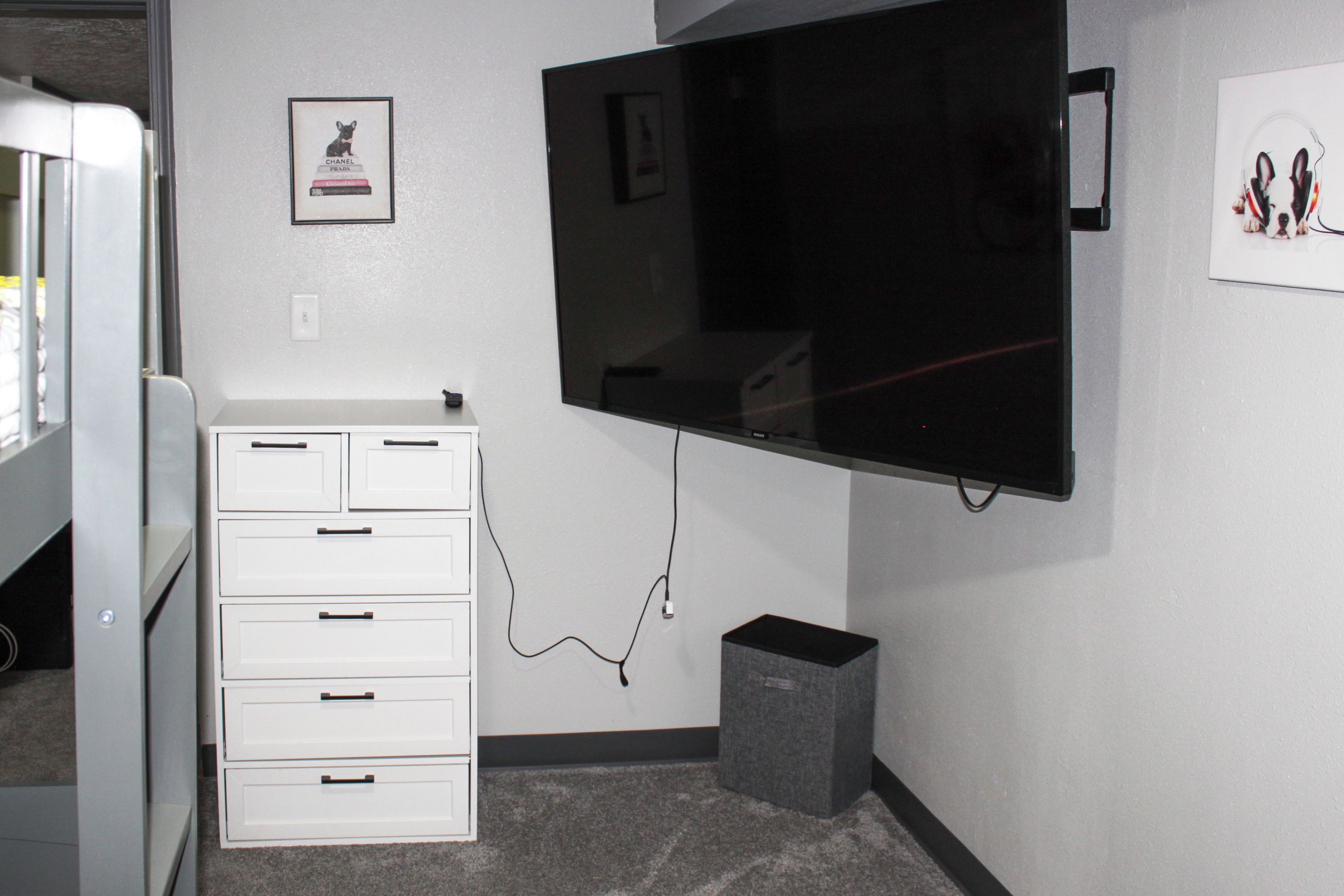 A room with a flat-screen TV mounted on the wall, a white dresser with drawers, and a small gray speaker on the floor.