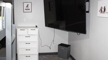 A room with a flat-screen TV mounted on the wall, a white dresser with drawers, and a small gray speaker on the floor.