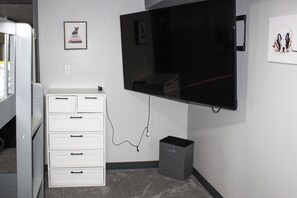 A room with a flat-screen TV mounted on the wall, a white dresser with drawers, and a small gray speaker on the floor.