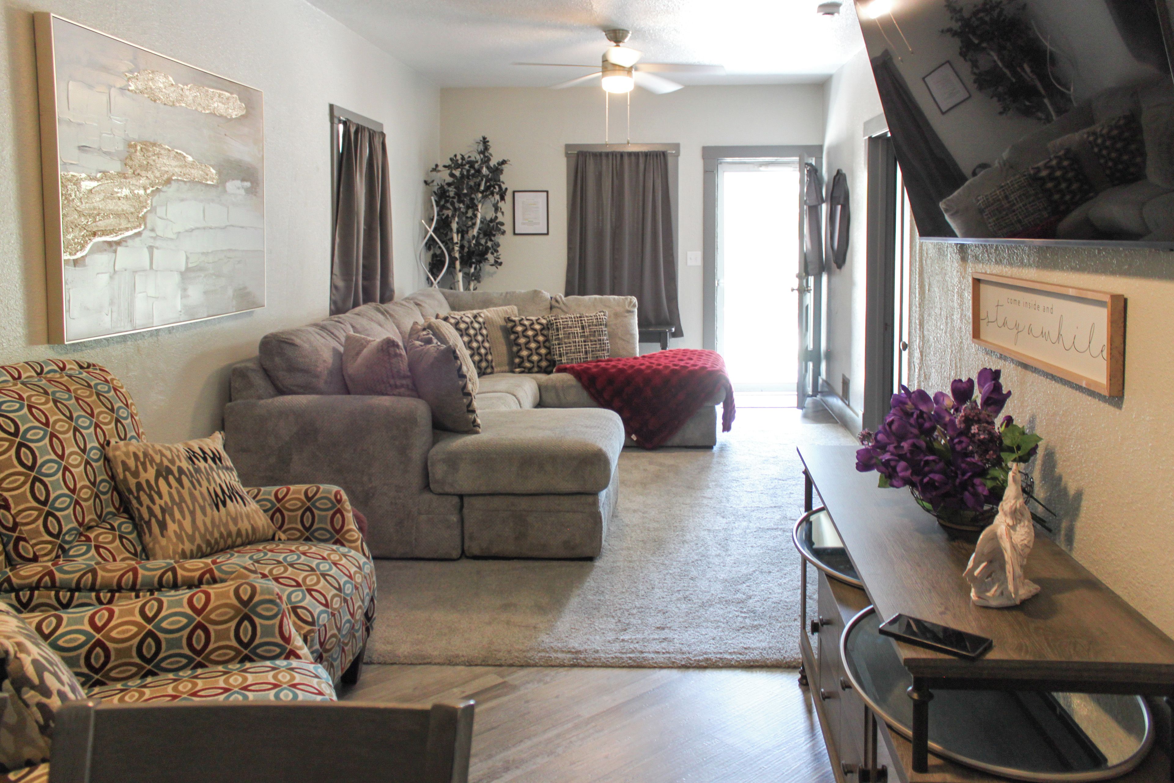 A living room with a sofa, a patterned armchair, a dining table with a vase of flowers, and a ceiling fan.