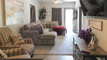 A living room with a sofa, a patterned armchair, a dining table with a vase of flowers, and a ceiling fan.