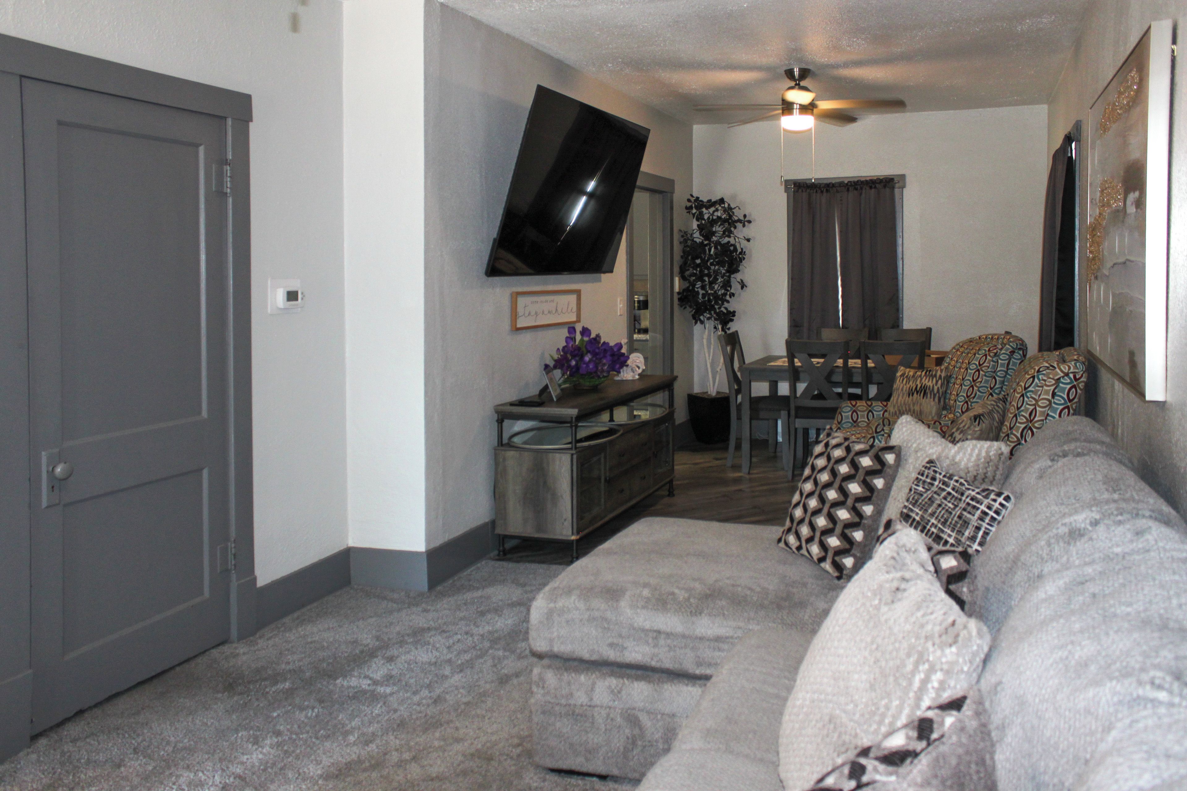 A living room with a gray couch, a flat-screen TV mounted on the wall, a ceiling fan, and a dining area with a table and chairs.
