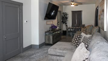 A living room with a gray couch, a flat-screen TV mounted on the wall, a ceiling fan, and a dining area with a table and chairs.