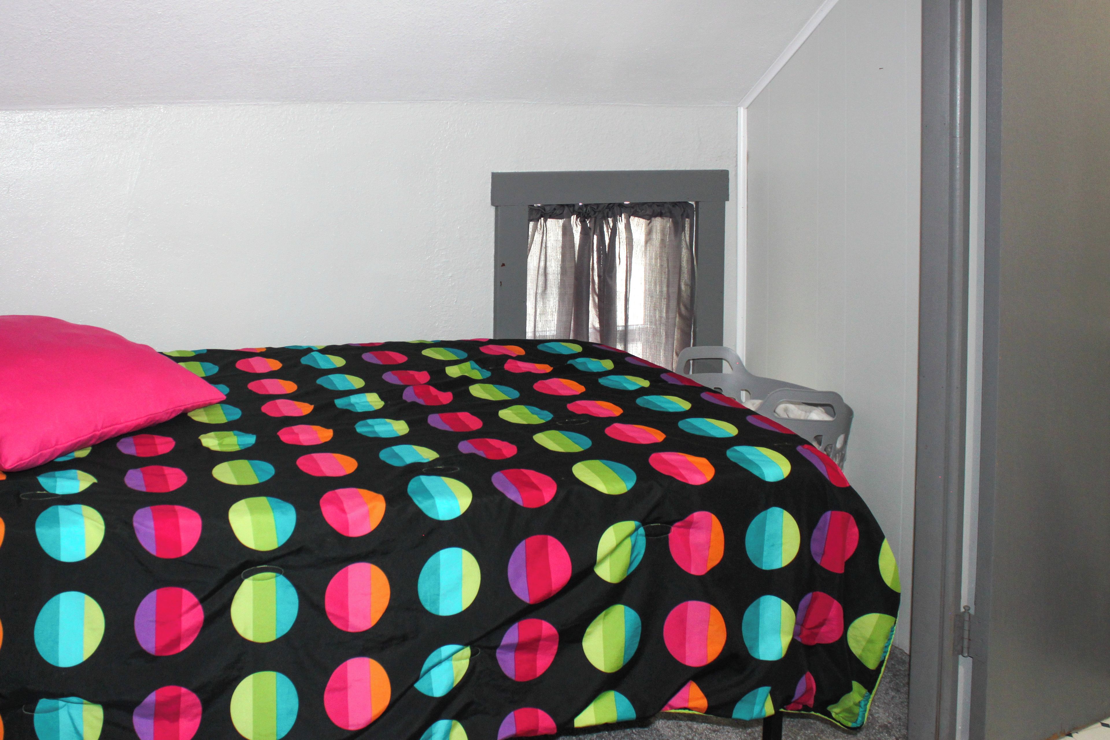 Quarto com uma cama coberta por um cobertor estampado em bolinhas, uma janela com cortinas e uma mesa de cabeceira.
