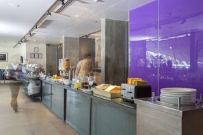 A modern breakfast bar with a glass partition, a buffet area with food items, and a view of a cityscape through a large window. - Mana Hotel Ranakpur (Desuri)