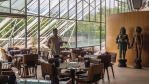 A restaurant with a high ceiling and large windows, featuring a waiter and two statues on the wall. - Mana Hotel Ranakpur (Desuri)