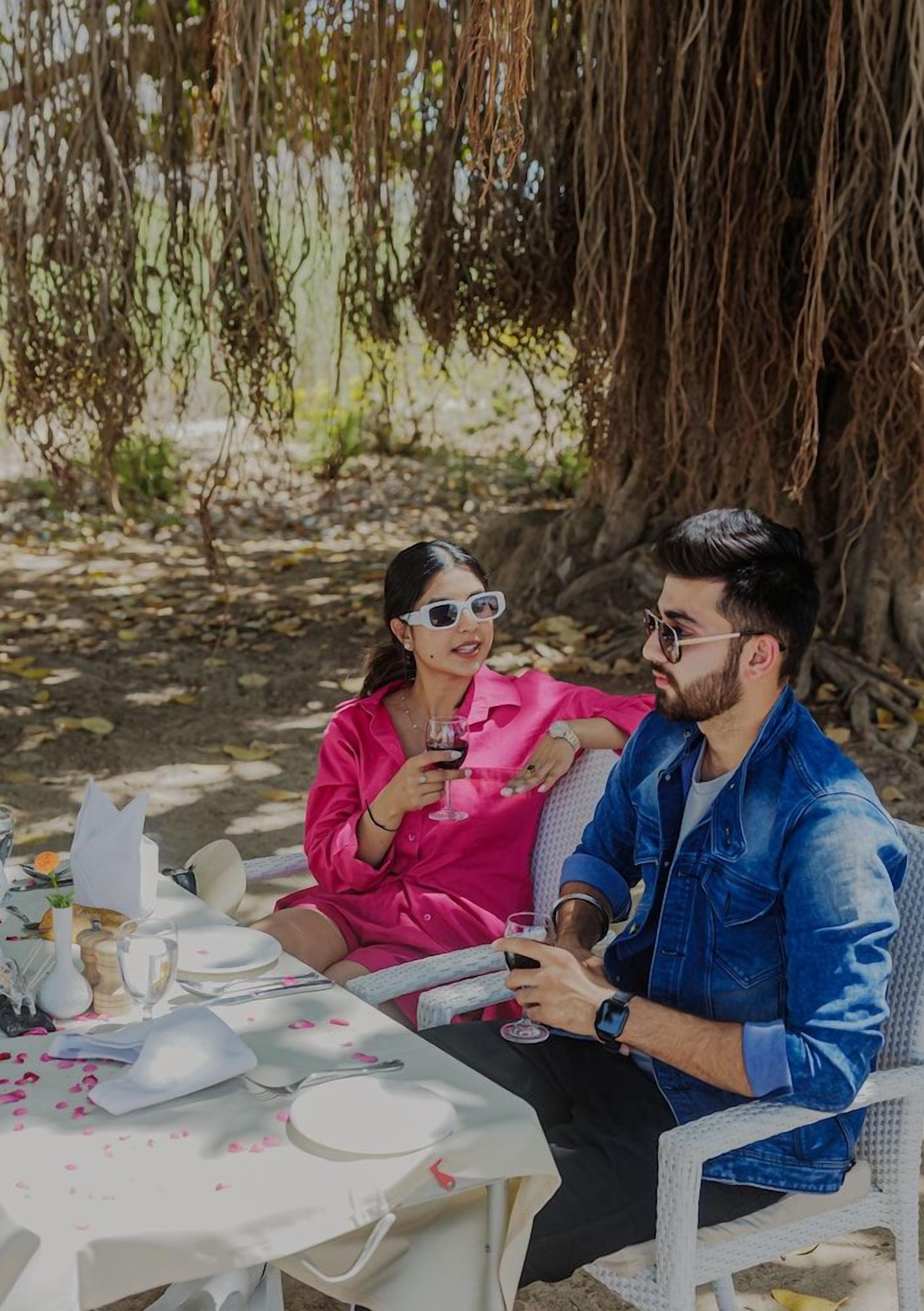 Dos personas sentadas en una mesa al aire libre con mantel blanco, rodeadas de follaje y un árbol.