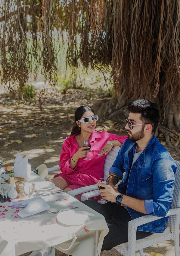 Two people seated at an outdoor table with a white tablecloth, surrounded by greenery and a tree.