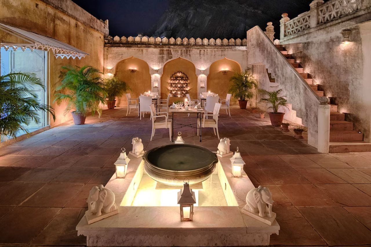An outdoor dining area with a fountain centerpiece, surrounded by potted plants and a staircase leading to an upper level.