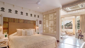 A spacious bedroom with a large bed, a nightstand, a decorative wall, and a view through a window. - Rawla Narlai Luxury Heritage Hotel (Desuri)