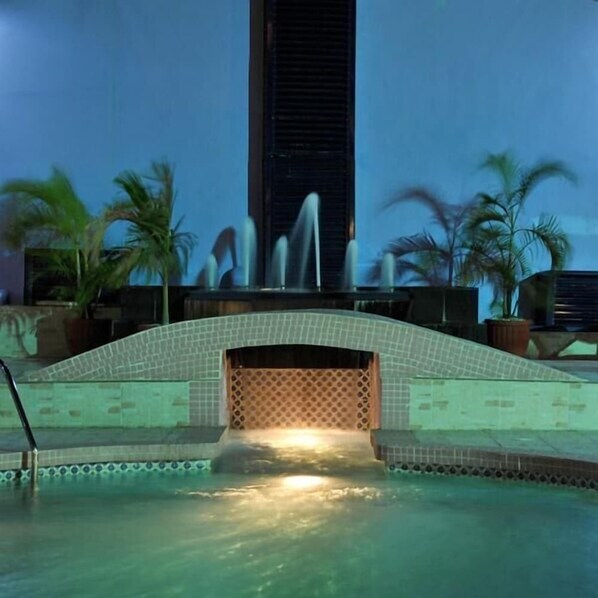 Un área de piscina con bordeado de azulejos, palmeras y una fuente de agua.