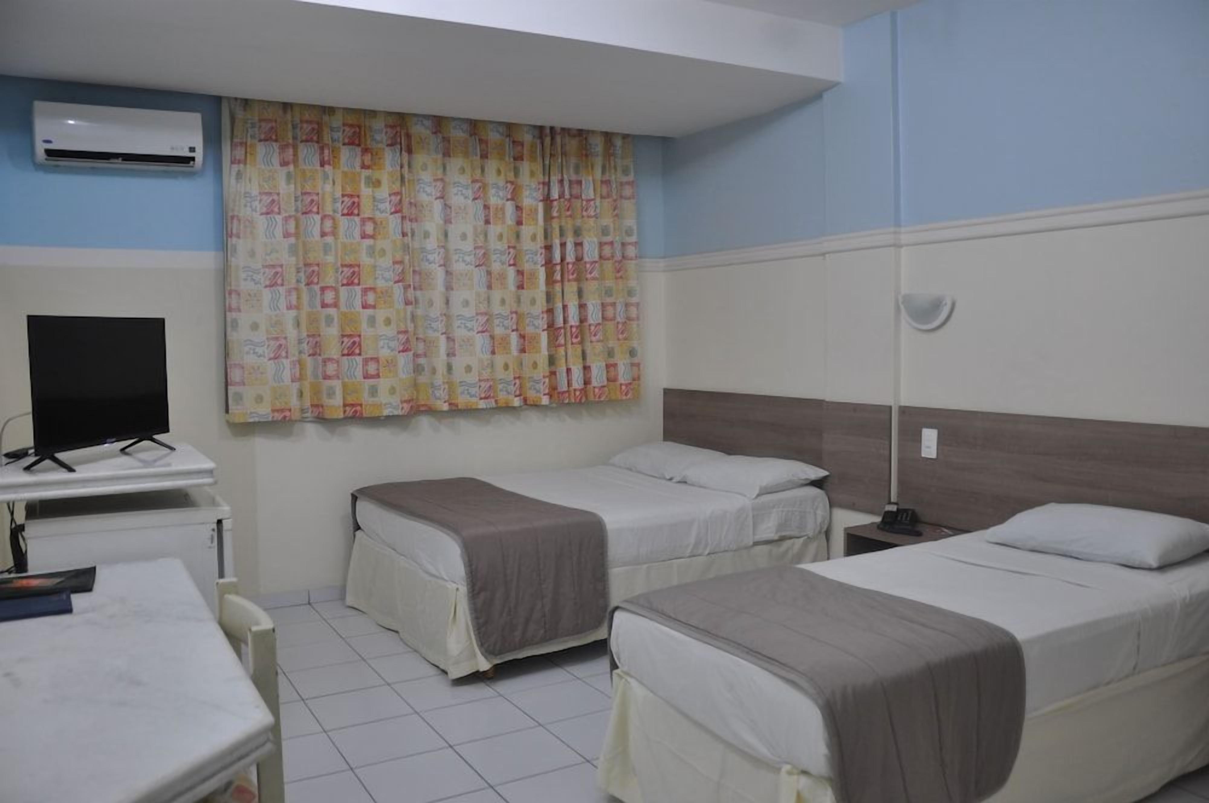 Quarto de hotel com duas camas de solteiro, uma escrivaninha com televisão de tela plana, mesa de cabeceira e cortina estampada.