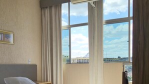 A hotel room with a bed, a desk, and a large window with curtains. - Real Minas Suites Hotel (Governador Valadares)