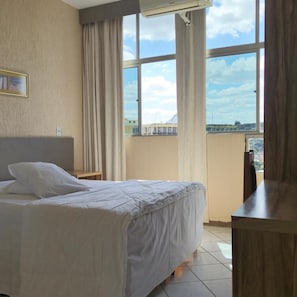 A hotel room with a bed, a desk, and a large window with curtains. - Real Minas Suites Hotel (Governador Valadares)