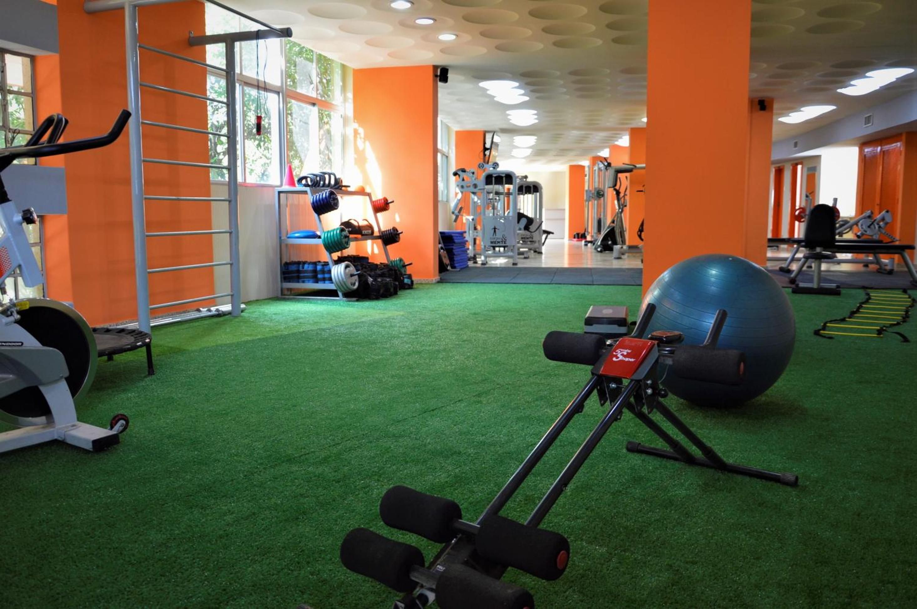 Academia com equipamentos de ginástica, grama sintética verde e paredes laranja.